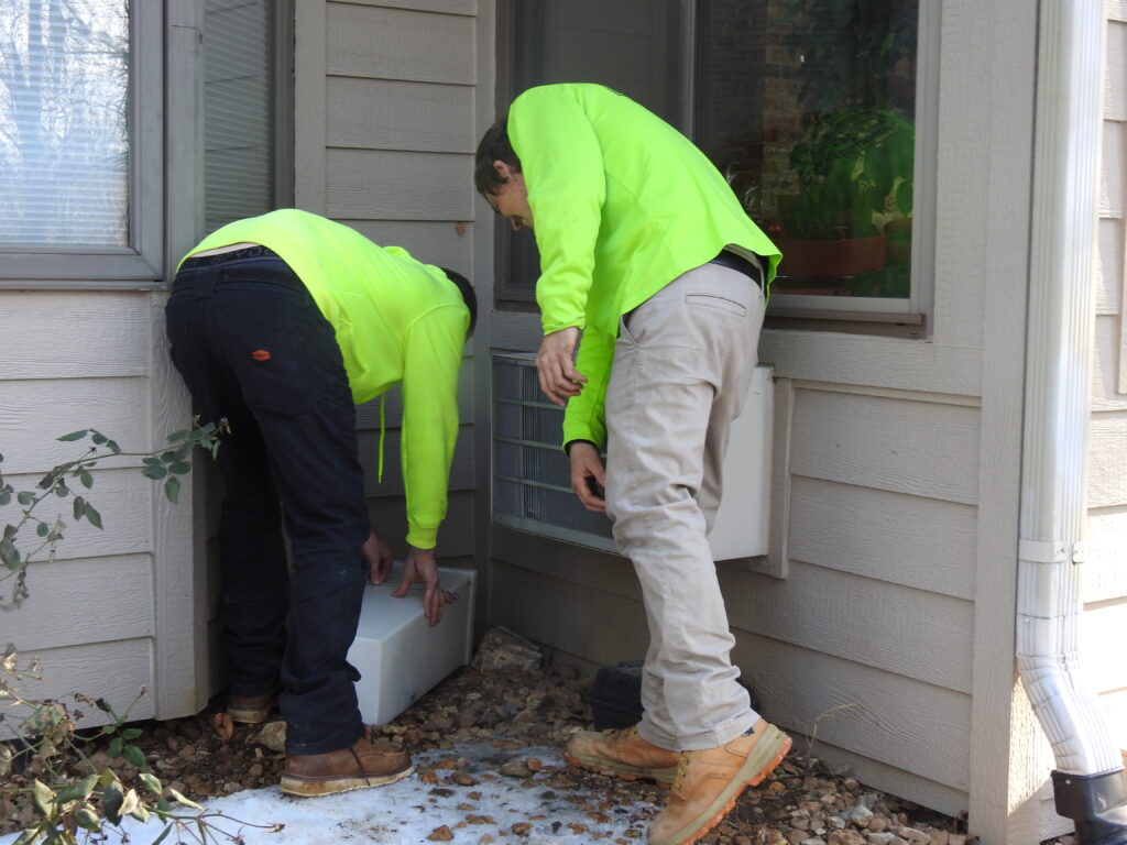 Pest control technicians installing a rodent bait station near a home's foundation in Pacific, MO (63069).