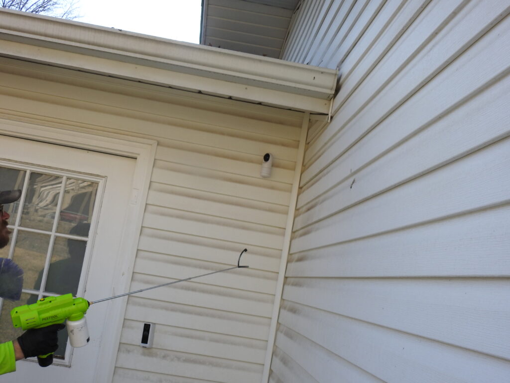 Pest control technician applying pest treatment to exterior siding near a door and security camera in St. Peters, MO 63376.