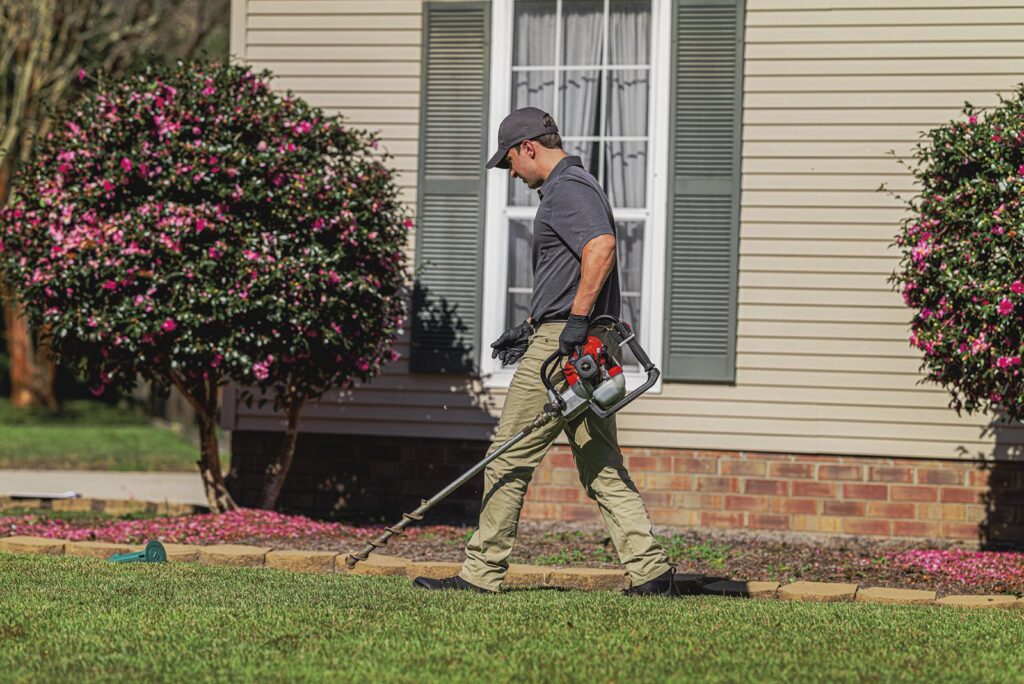 A pest control technician applying a targeted treatment around the exterior foundation of a landscaped home